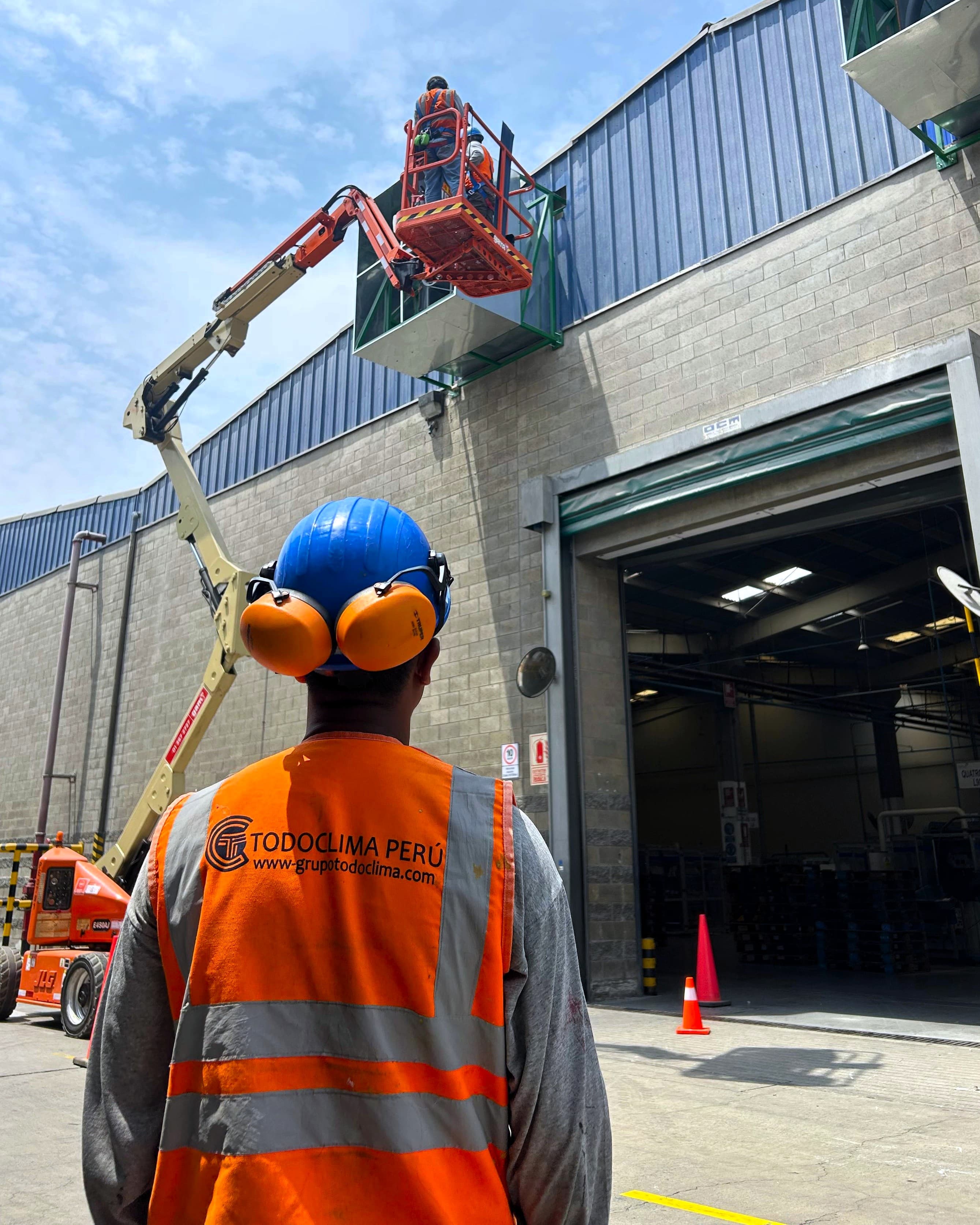 Técnico de Grupo TodoClima con chaleco y casco observa a sus compañeros trabajar desde una plataforma elevadora sobre la nave industrial, junto a los equipos de climatización instalados en la cubierta.